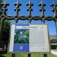 Übersicht Schloss Fürstenberg Informationstafel vor einem Eisengitter, dahinter Schlossbauten und blauer Himmel sichtbar.