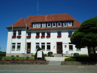 Das weiße, zweistöckige Rathaus von Bad Wünnenberg mit roten Dachziegeln und blühenden Blumenkästen.