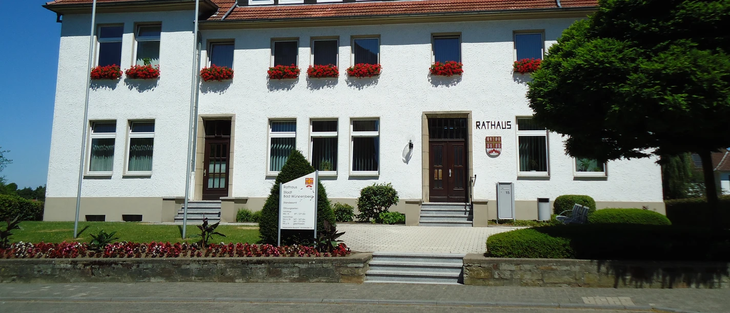Das weiße, zweistöckige Rathaus von Bad Wünnenberg mit roten Dachziegeln und blühenden Blumenkästen.