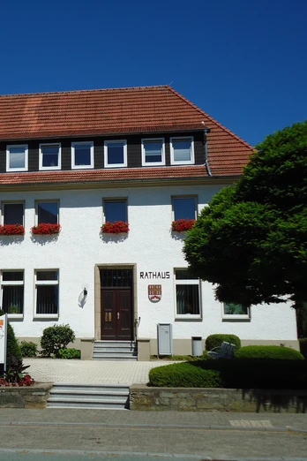 Das weiße, zweistöckige Rathaus von Bad Wünnenberg mit roten Dachziegeln und blühenden Blumenkästen.