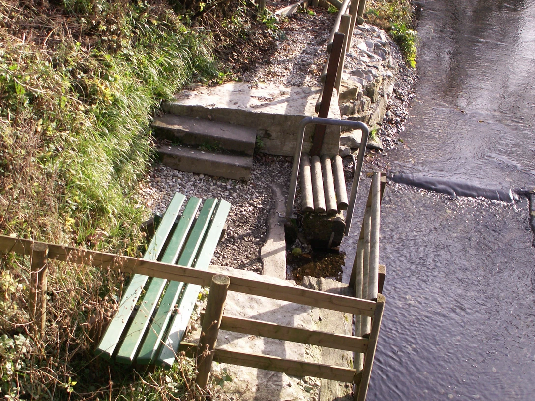 Flusstretbecken mit grünem Geländer, Stufen und Sitzbank am Bachlauf, umgeben von Natur.
