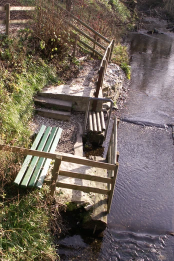 Flusstretbecken mit grünem Geländer, Stufen und Sitzbank am Bachlauf, umgeben von Natur.