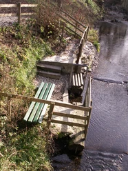 Flusstretbecken Frei- und Hallenbad Flusstretbecken mit grünem Geländer, Stufen und Sitzbank am Bachlauf, umgeben von Natur.