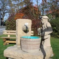 Eine lebensgroße Steinskulptur eines Mannes, der Wasser in ein Fass gießt, umgeben von grüner Natur.