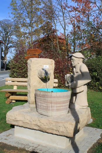 Eine lebensgroße Steinskulptur eines Mannes, der Wasser in ein Fass gießt, umgeben von grüner Natur.