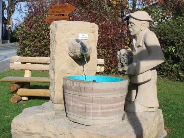 Bleiwäscher Skulptur Eine lebensgroße Steinskulptur eines Mannes, der Wasser in ein Fass gießt, umgeben von grüner Natur.