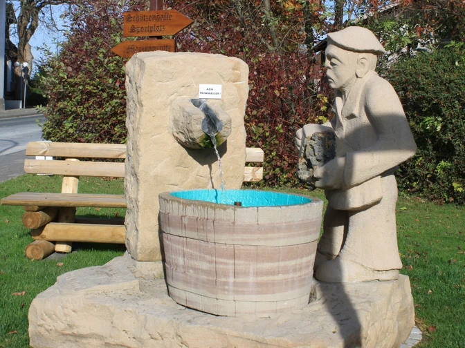 Bleiwäscher Skulptur Eine lebensgroße Steinskulptur eines Mannes, der Wasser in ein Fass gießt, umgeben von grüner Natur.