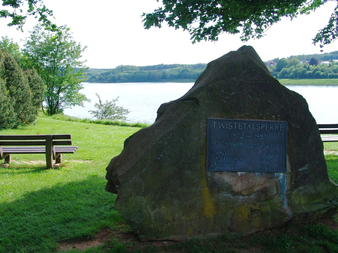 Gedenkstein der Twistetalsperre am Ufer eines Sees, umgeben von Bäumen und Parkbänken im Grün.