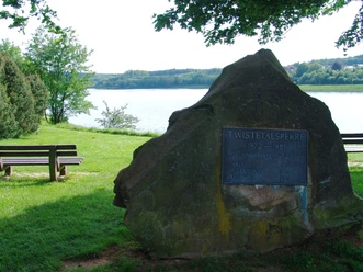 Twistetalsperre Gedenkstein der Twistetalsperre am Ufer eines Sees, umgeben von Bäumen und Parkbänken im Grün.