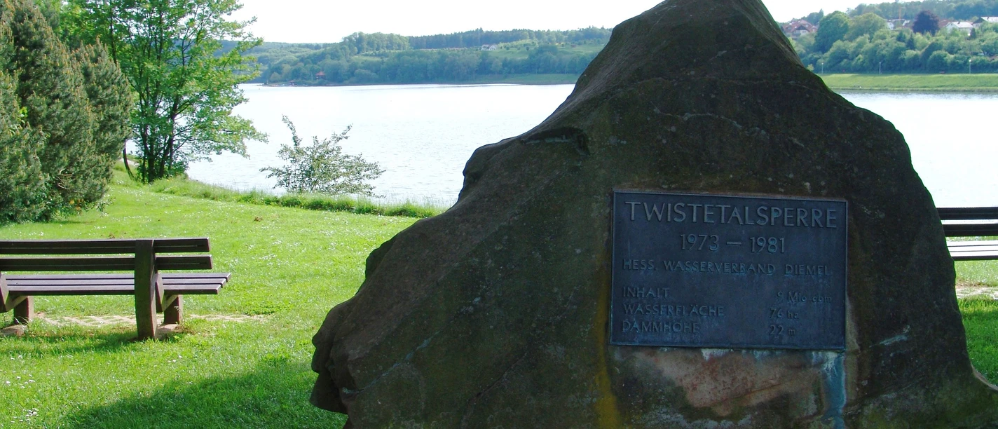 Twistetalsperre Gedenkstein der Twistetalsperre am Ufer eines Sees, umgeben von Bäumen und Parkbänken im Grün.