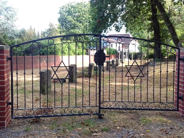 Eisenzaun mit Davidstern-Motiven führt zu einem historischen Friedhof, gesäumt von alten Grabsteinen.
