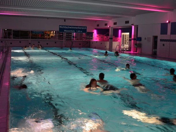 Schwimmbecken mit abendlicher Beleuchtung, Bademöglichkeiten und einige Menschen beim Schwimmen.