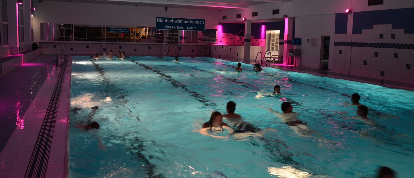 Hallenbad in Brakel Schwimmbecken mit abendlicher Beleuchtung, Bademöglichkeiten und einige Menschen beim Schwimmen.
