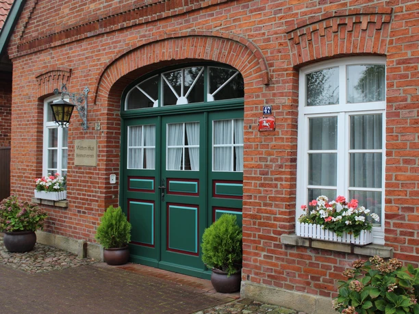 Witten Hus Grüner Holzeingang und weiße Fenster eines Backsteingebäudes, flankiert von blühenden Pflanzen.
