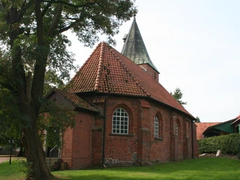 Backsteinkirche mit rotem Ziegeldach, an einem Baum gelegen, eingefasst von einem gepflegten Rasen.