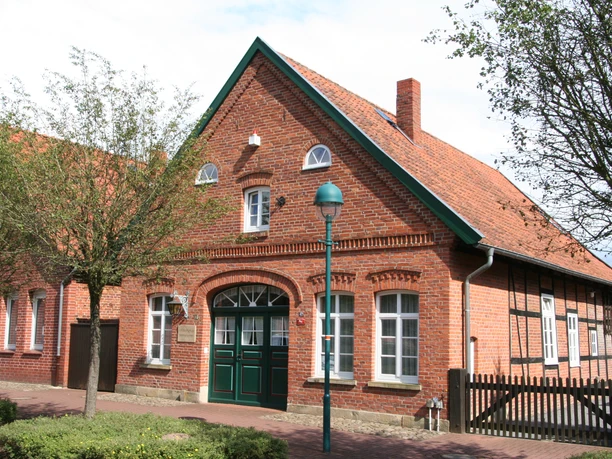 Brick building with gabled roof and green doors, decorated with white arches, trees and a lantern in front.