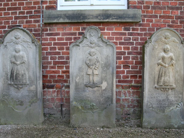 Drei historische Grabsteine aus Stein mit Reliefs und Inschriften an roter Backsteinwand.