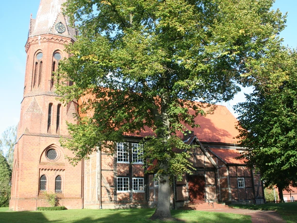Ev.-luth. Kirche zu Estorf Ein roter Backsteinbau der kirchlichen Architektur mit spitzem Turm und grünen Bäumen im Vordergrund.