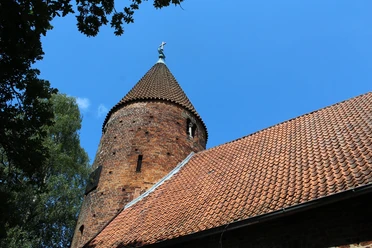 Brick church with striking round tower and slate roof, surrounded by trees and blue sky.