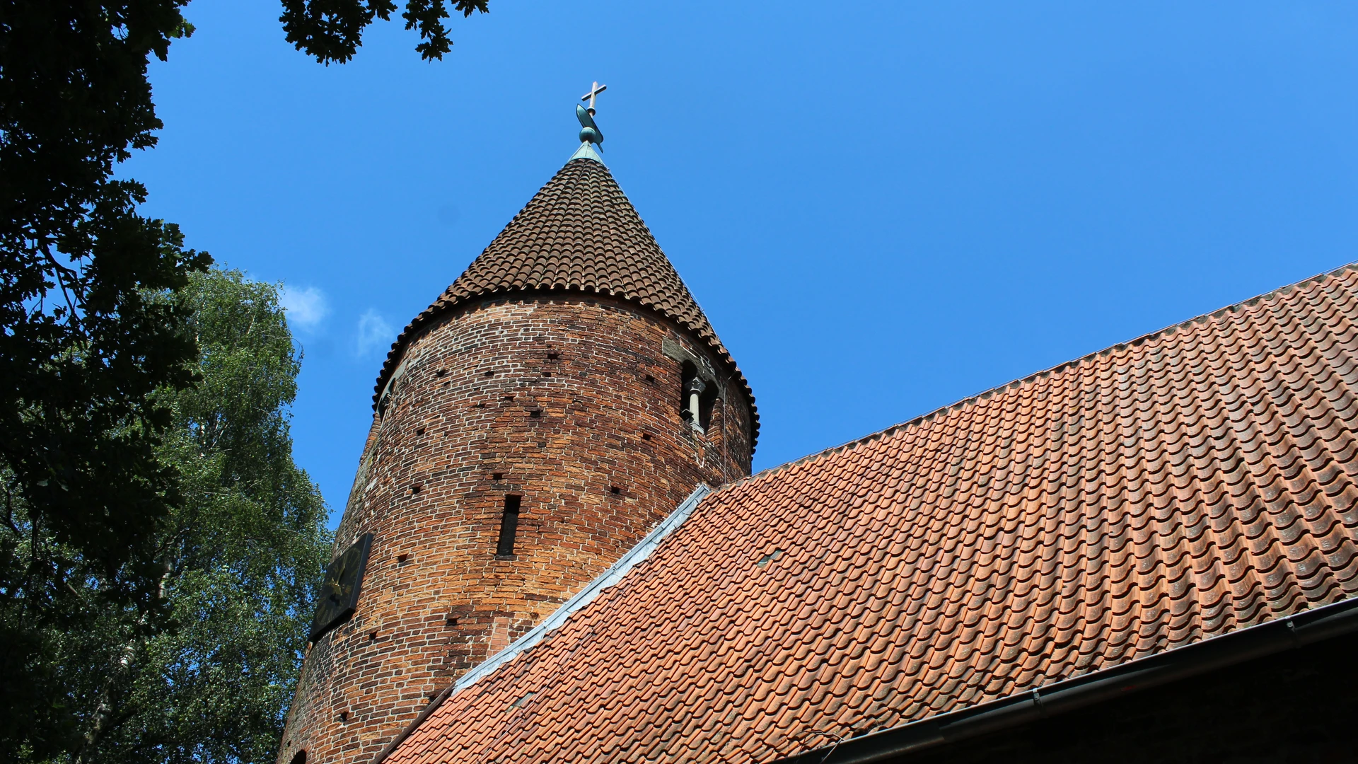 Brick church with striking round tower and slate roof, surrounded by trees and blue sky.