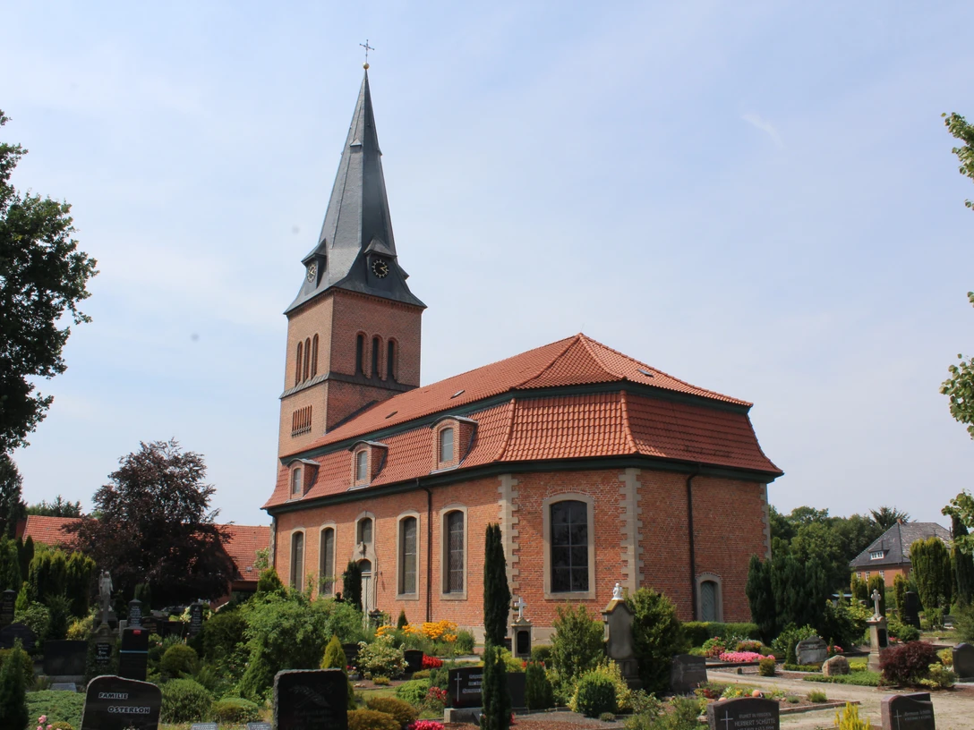 Backsteinkirche mit spitzem Turm vor einem blauen Himmel, umgeben von einem grünen Friedhof.