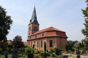 Kirche Schwarme Backsteinkirche mit spitzem Turm vor einem blauen Himmel, umgeben von einem grünen Friedhof.