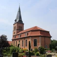 Kirche Schwarme Backsteinkirche mit spitzem Turm vor einem blauen Himmel, umgeben von einem grünen Friedhof.