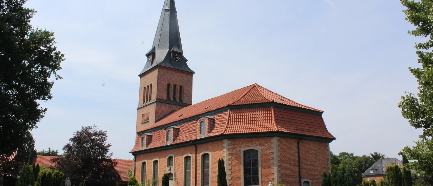 Kirche Schwarme Backsteinkirche mit spitzem Turm vor einem blauen Himmel, umgeben von einem grünen Friedhof.