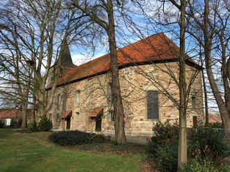 Historische Steinkirche mit rotem Ziegeldach und Turm, umgeben von kahlen Bäumen an einem klaren Tag.