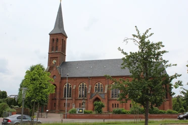 Backsteinkirche mit hohem Spitzturm und grünen Bäumen, die majestätisch in einem ruhigen Umfeld steht.