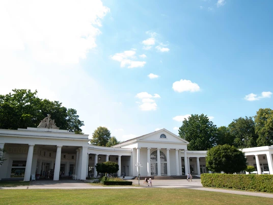 Wandelhalle © Tourismus NRW.JPG Das Bild zeigt die Außenansicht der Wandelhalle mit einer weißen, historischen Fassade. Kleine Bäume umgeben das Gebäude, das mitten im Kurpark steht.