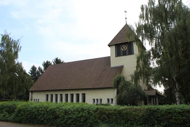 Kirche in Essern mit markantem Glockenturm, umgeben von Bäumen und von einer Hecke eingefasst.