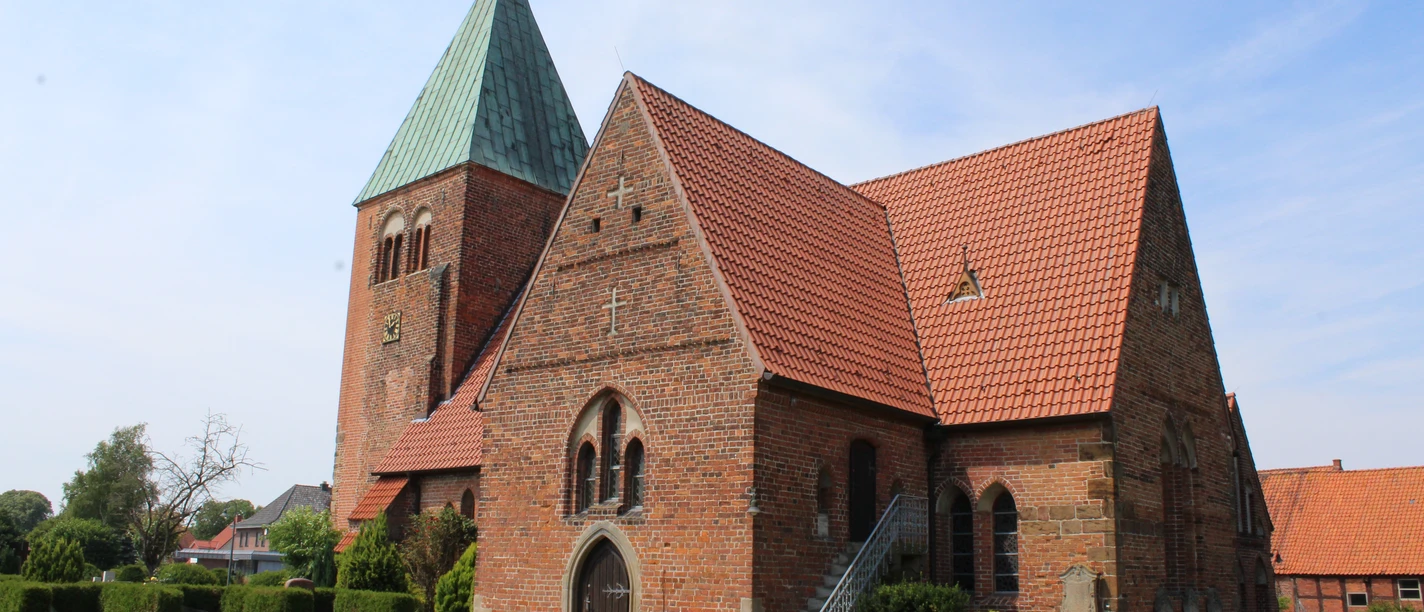 Backsteinkirche mit grünem Turmdach, Satteldächern und gepflegtem Grün vor blauem Himmel.