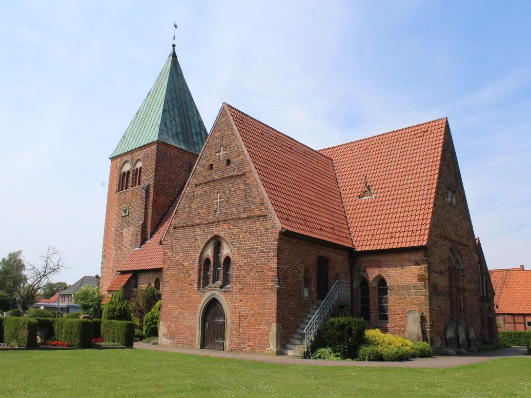 Kirche Riede Backsteinkirche mit grünem Turmdach, Satteldächern und gepflegtem Grün vor blauem Himmel.
