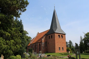 Backsteinkirche St. Crucis in Hoyerhagen mit spitzem Kirchturm umgeben von Bäumen und einem Friedhof.