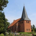 Backsteinkirche St. Crucis in Hoyerhagen mit spitzem Kirchturm umgeben von Bäumen und einem Friedhof.