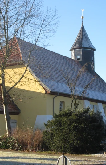 Historische Kirche mit gelbem Anstrich und markantem Holzturm, umgeben von Bäumen im Winter.