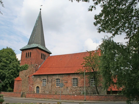 Kirche Holtorf Backsteinkirche mit markantem Spitzdach, umgeben von Bäumen, vor bewölktem Himmel in Holtorf.Brick church with a striking pointed roof, surrounded by trees, against a cloudy sky in Holtorf.Murstenskirke med et markant spidst tag, omgivet af træer, mod en overskyet himmel i Holtorf.Bakstenen kerk met een opvallend puntdak, omgeven door bomen, tegen een bewolkte lucht in Holtorf.