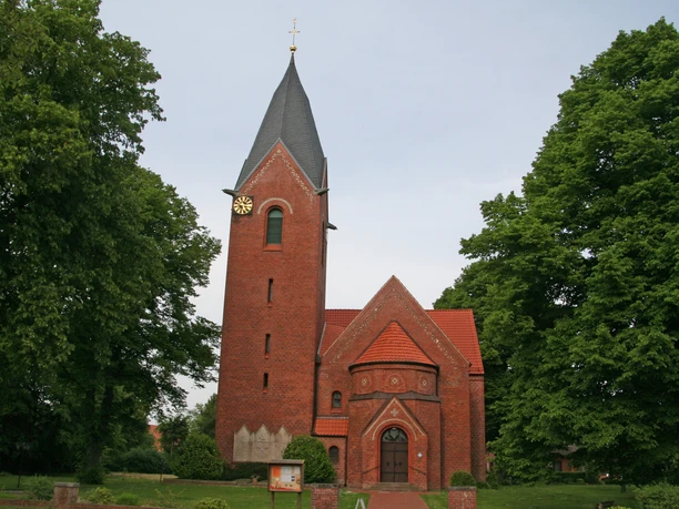 Alte Kapelle Schweringen Brick chapel with high tower and clock, surrounded by green trees under a cloudy sky.