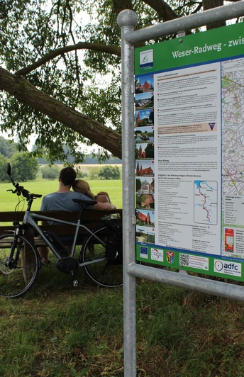 Zwei Radfahrer pausieren an einer Bank auf einer grünen Wiese, neben einem Info-Schild am Weser-Radweg.