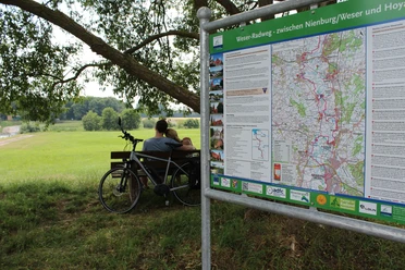 Radfahrer am Weser-Radweg Zwei Radfahrer pausieren an einer Bank auf einer grünen Wiese, neben einem Info-Schild am Weser-Radweg.