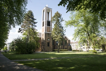 Auferstehungskirche am Kurpark, Bad Oeyhausen Auf dem Bild ist die Auferstehungskirche in Bad Oeynhausen zu sehen, umgeben von einer grünen Wiese. Die rot-braune Fassade der Kirche hebt sich gut ab, während Bäume das Bild rahmen und eine ruhige Atmosphäre schaffen.