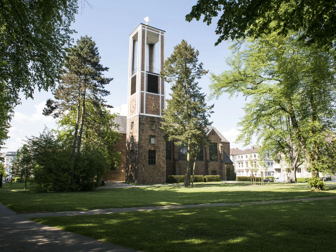 Auferstehungskirche am Kurpark, Bad Oeyhausen Auf dem Bild ist die Auferstehungskirche in Bad Oeynhausen zu sehen, umgeben von einer grünen Wiese. Die rot-braune Fassade der Kirche hebt sich gut ab, während Bäume das Bild rahmen und eine ruhige Atmosphäre schaffen.
