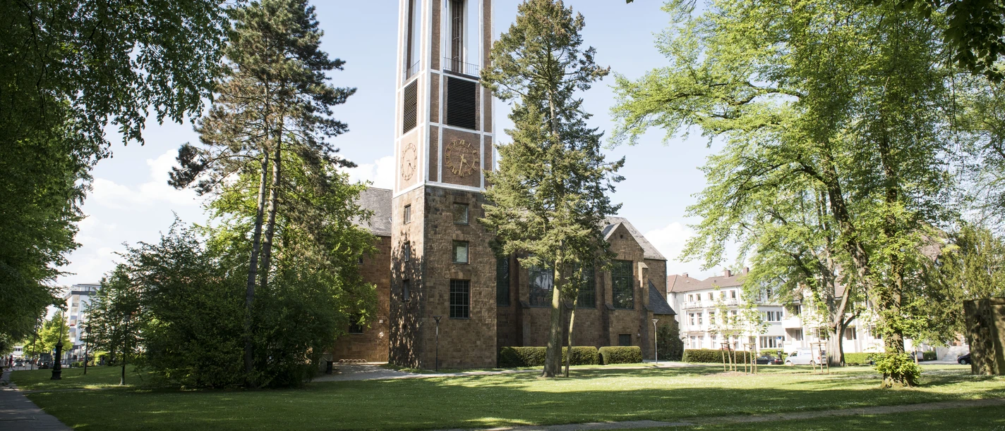 Auferstehungskirche am Kurpark, Bad Oeyhausen Auf dem Bild ist die Auferstehungskirche in Bad Oeynhausen zu sehen, umgeben von einer grünen Wiese. Die rot-braune Fassade der Kirche hebt sich gut ab, während Bäume das Bild rahmen und eine ruhige Atmosphäre schaffen.