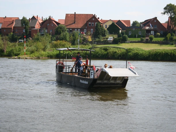 Ferry crossing river with houses in the background, passengers and the ferryman on board.
