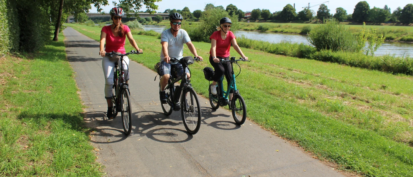 Three cyclists enjoy a sunny ride on a cycle path along the green Weser landscape.