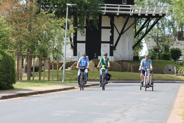 Three cyclists in sporty clothing ride along an asphalt road, with the Achim windmill in the background.