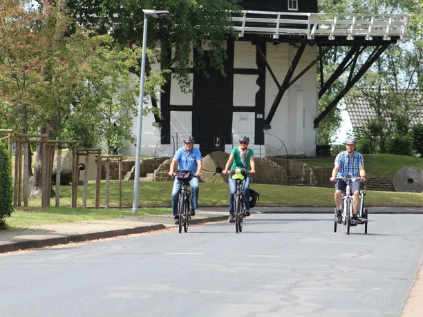 Three cyclists in sporty clothing ride along an asphalt road, with the Achim windmill in the background.