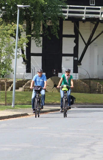 Radfahrer an der Achimer Windmühle Drei Radfahrer in sportlicher Kleidung fahren eine asphaltierte Straße entlang, im Hintergrund die Achimer Windmühle.