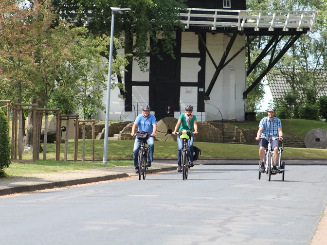 Radfahrer an der Achimer Windmühle Drei Radfahrer in sportlicher Kleidung fahren eine asphaltierte Straße entlang, im Hintergrund die Achimer Windmühle.Three cyclists in sporty clothing ride along an asphalt road, with the Achim windmill in the background.Tre cyklister i sportstøj kører ad en asfalteret vej med Achim-vindmøllen i baggrunden.Drie fietsers in sportieve kleding rijden over een geasfalteerde weg, met de Achim windmolen op de achtergrond.